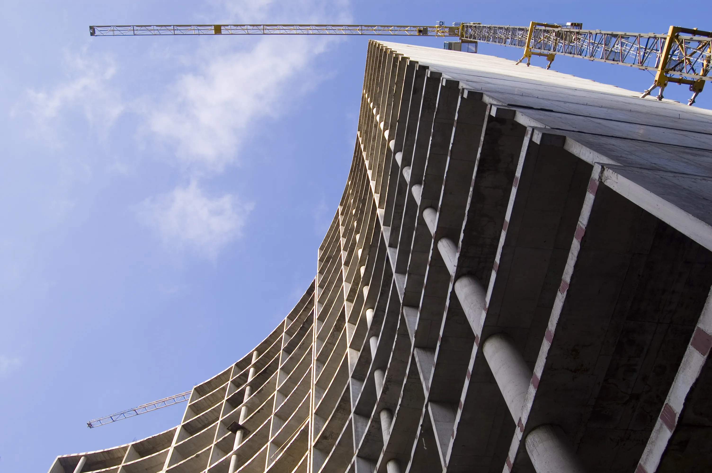 Low-angle view of a curved concrete building under construction with cranes against a blue sky.