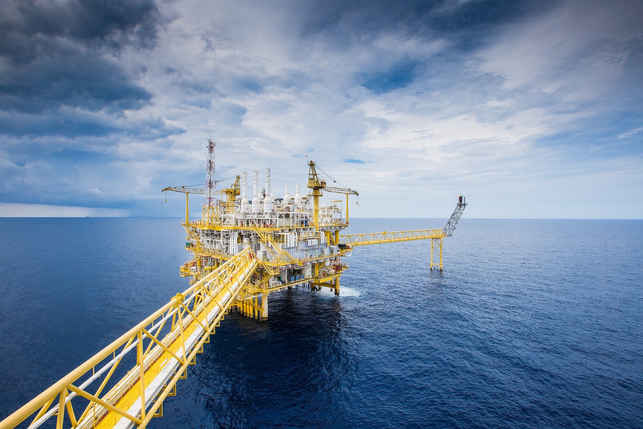 Offshore oil platform in the ocean with yellow metal walkways and cranes under a cloudy sky
