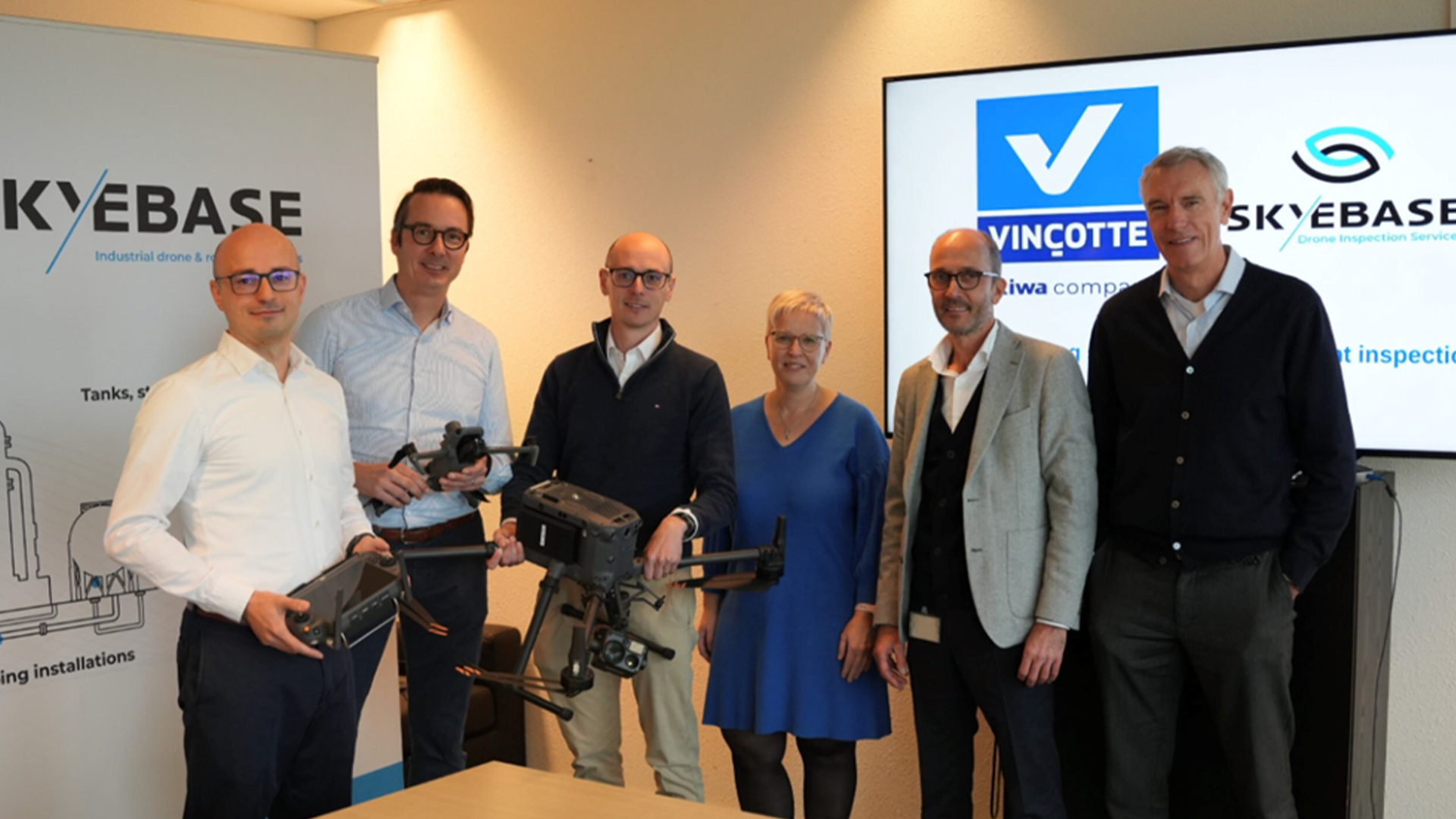 Group of six people standing indoors with industrial drone equipment, in front of Skybase and Vinçotte branded display screens.