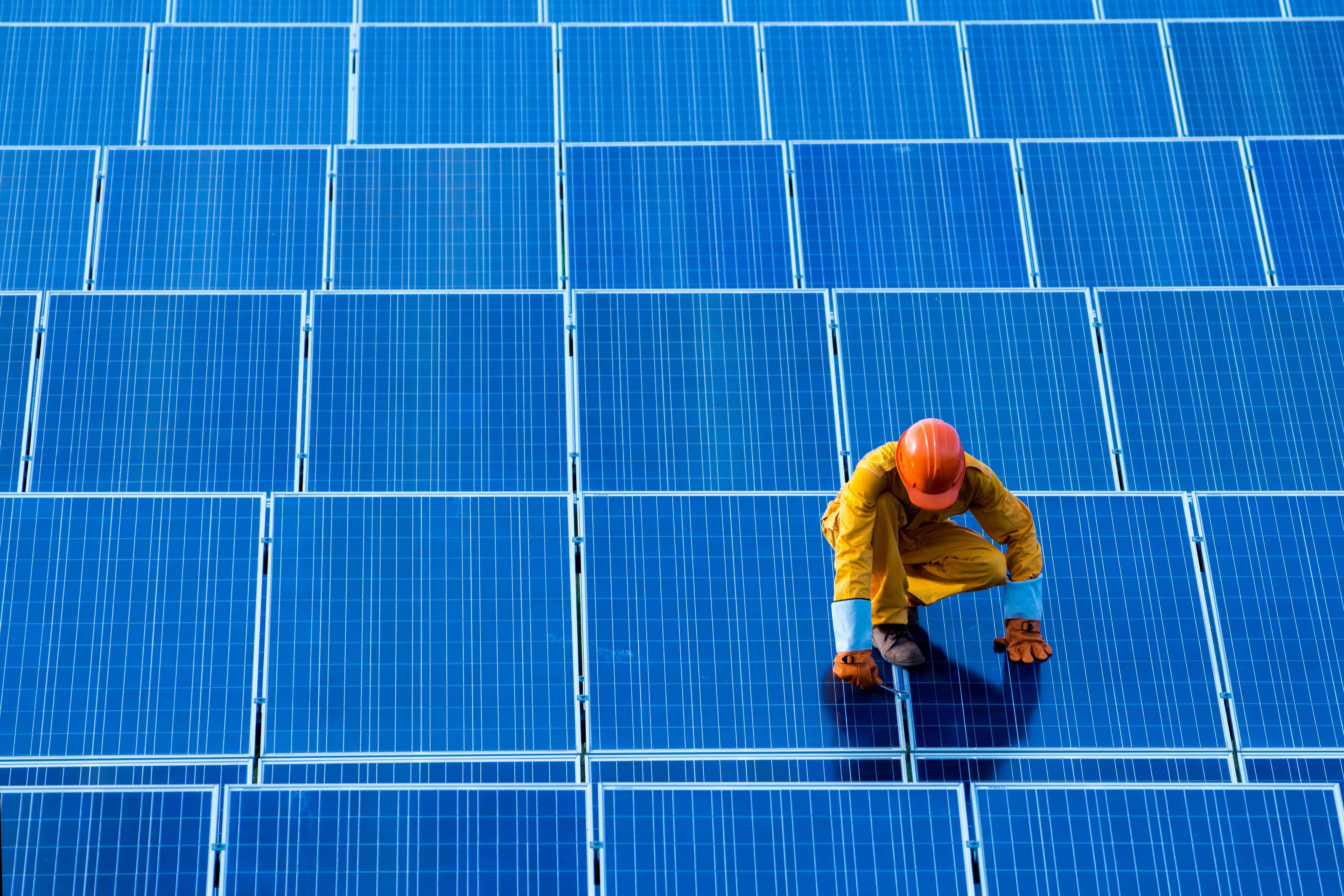 PV worker in safety gear inspecting rows of blue solar panels on a rooftop.