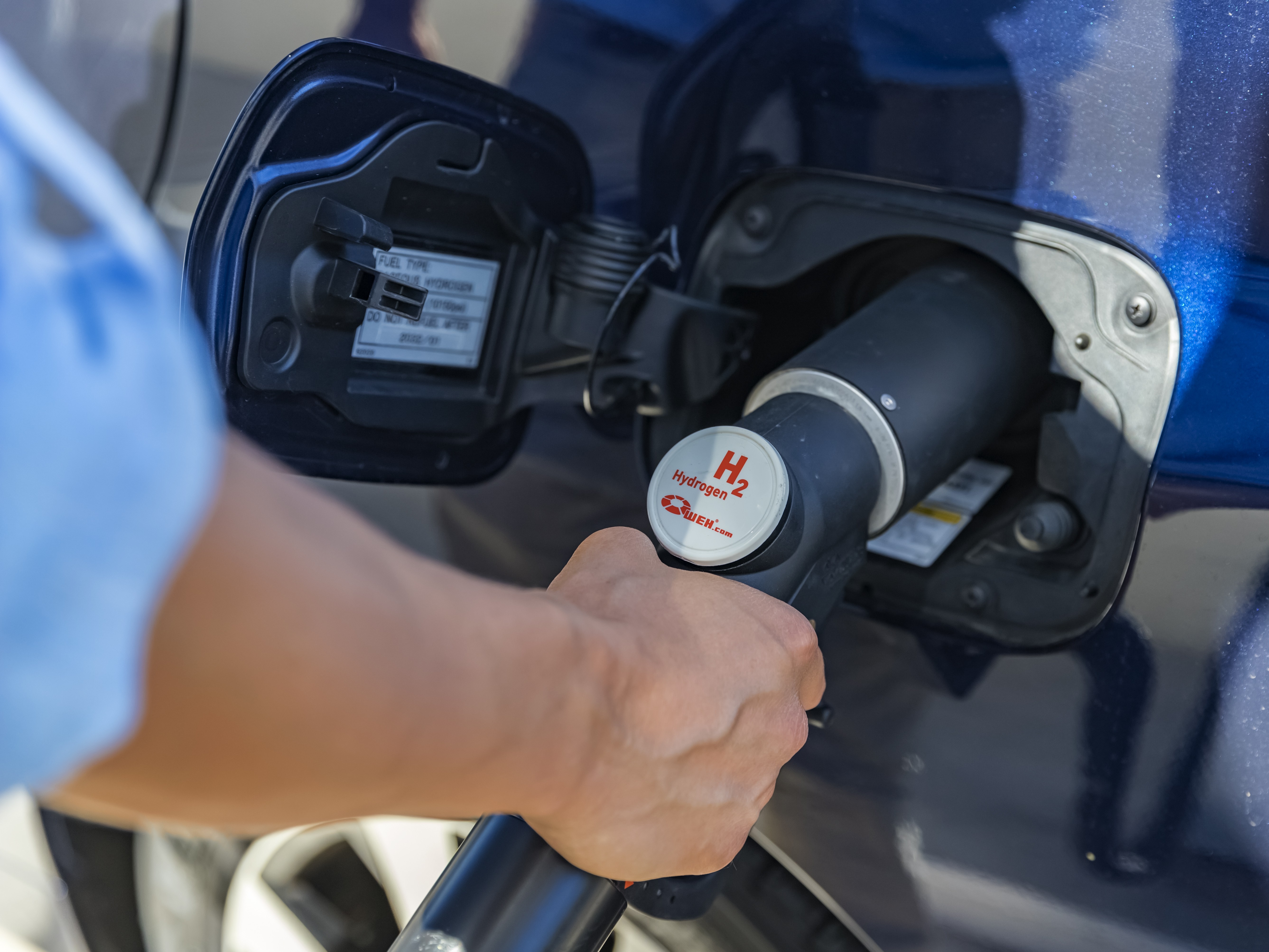 Close-up of a person refueling a blue vehicle with a hydrogen pump nozzle at a fuel port.