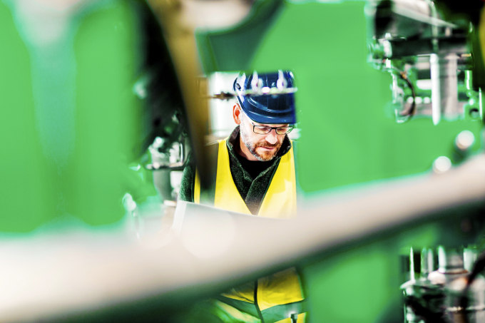 A Kiwa inspector wearing a hard hat and high-visibility vest inspects machinery in an industrial setting, framed by blurred green equipment in the foreground