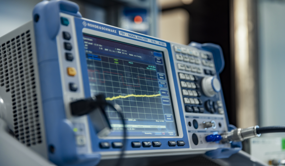 Close-up of a Rohde & Schwarz signal analyzer displaying a waveform graph with connected test cables and control buttons.