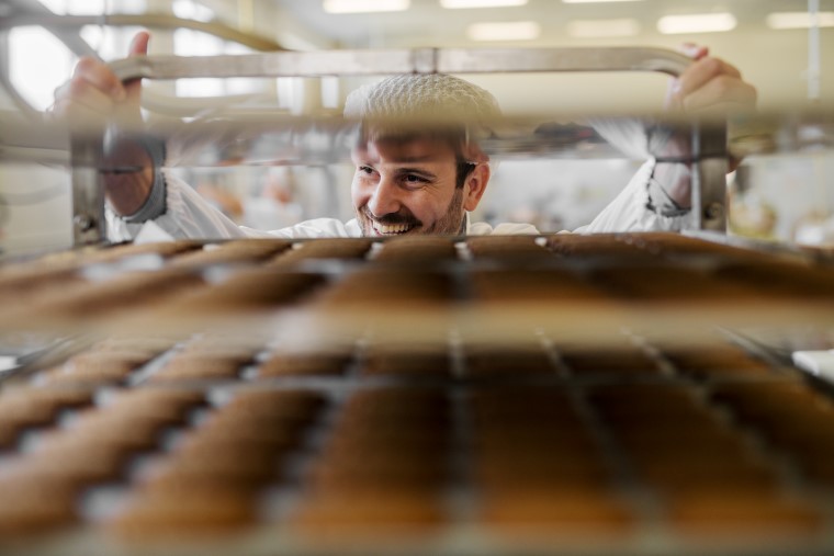 Baker in a hairnet and white coat handling a rack of baked goods in a commercial kitchen.