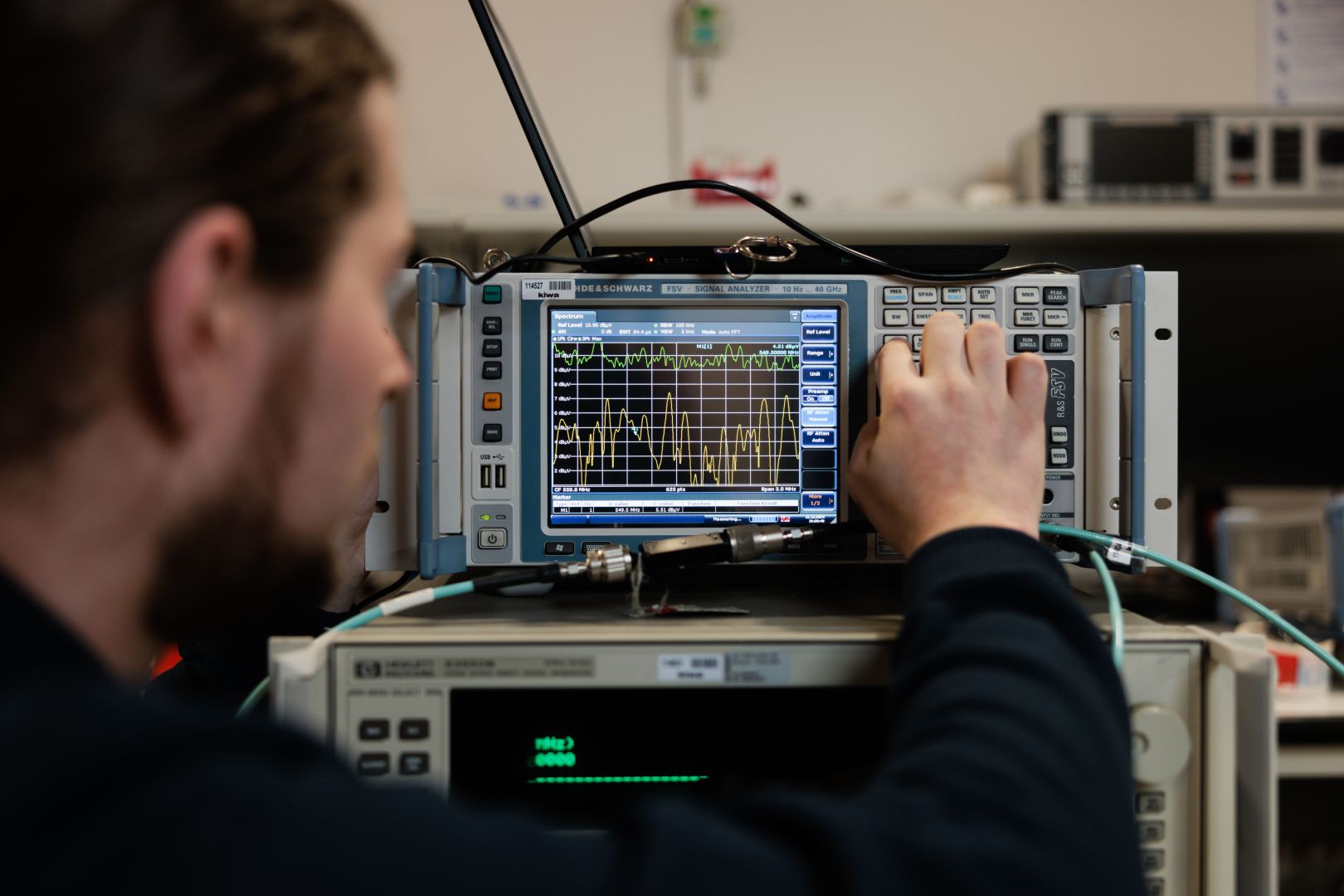 Kiwa technician adjusting a spectrum analyzer in an electronics lab, focusing on detailed waveform patterns displayed on the screen.
