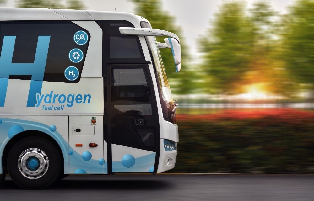 Side view of a hydrogen fuel cell bus driving on a road, with motion-blurred trees and sunlight in the background.