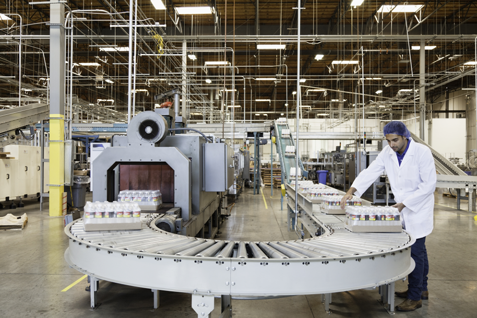 Worker in a white lab coat and hairnet organizing cans on a conveyor belt in a modern food processing plant.