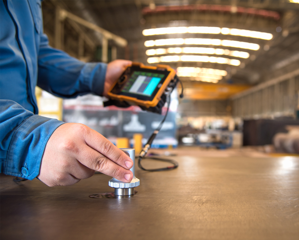 Kiwa laboratory employee using handheld device for ultrasonic testing in industrial setting.