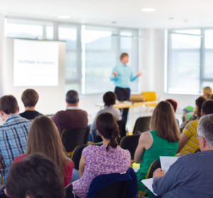 Audience attending a business presentation in a bright conference room, featuring a speaker in front of a projection screen.