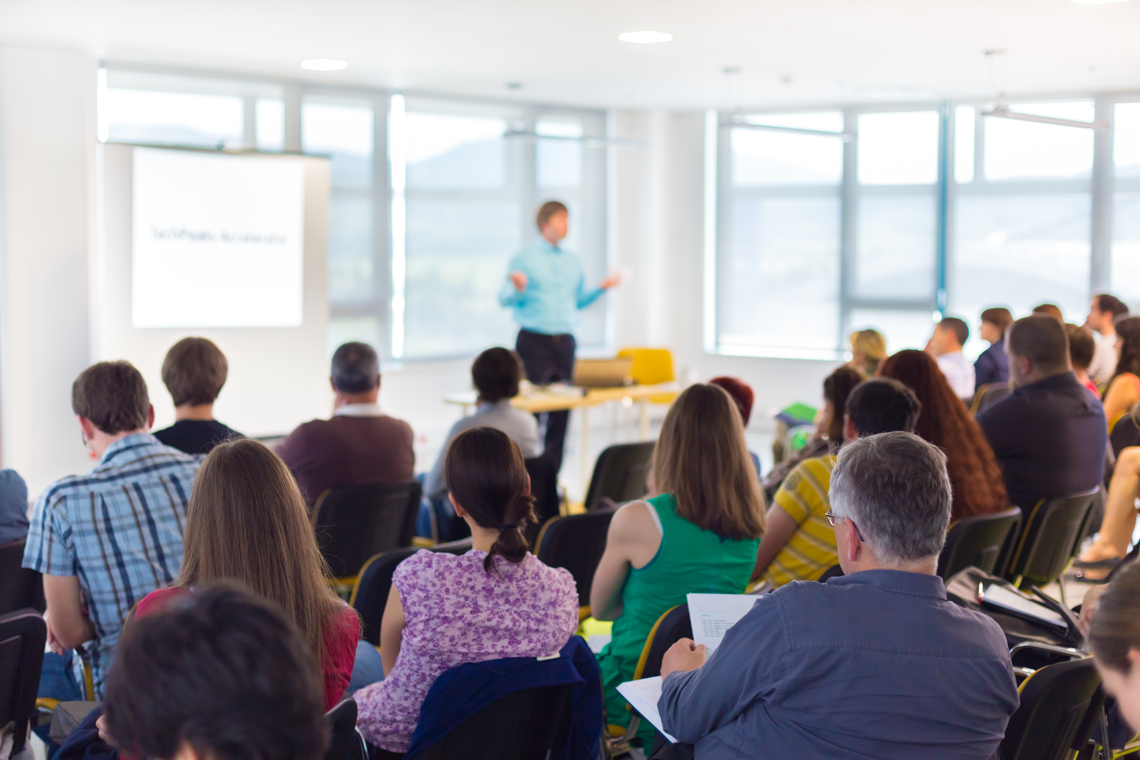 Audience attending a business presentation in a bright conference room, with a speaker standing near a projected screen.