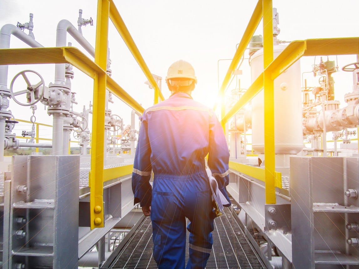 Kiwa PPI engineer in blue protective gear walking through an industrial oil and gas plant platform, surrounded by pipelines and machinery, with sunlight in the background
