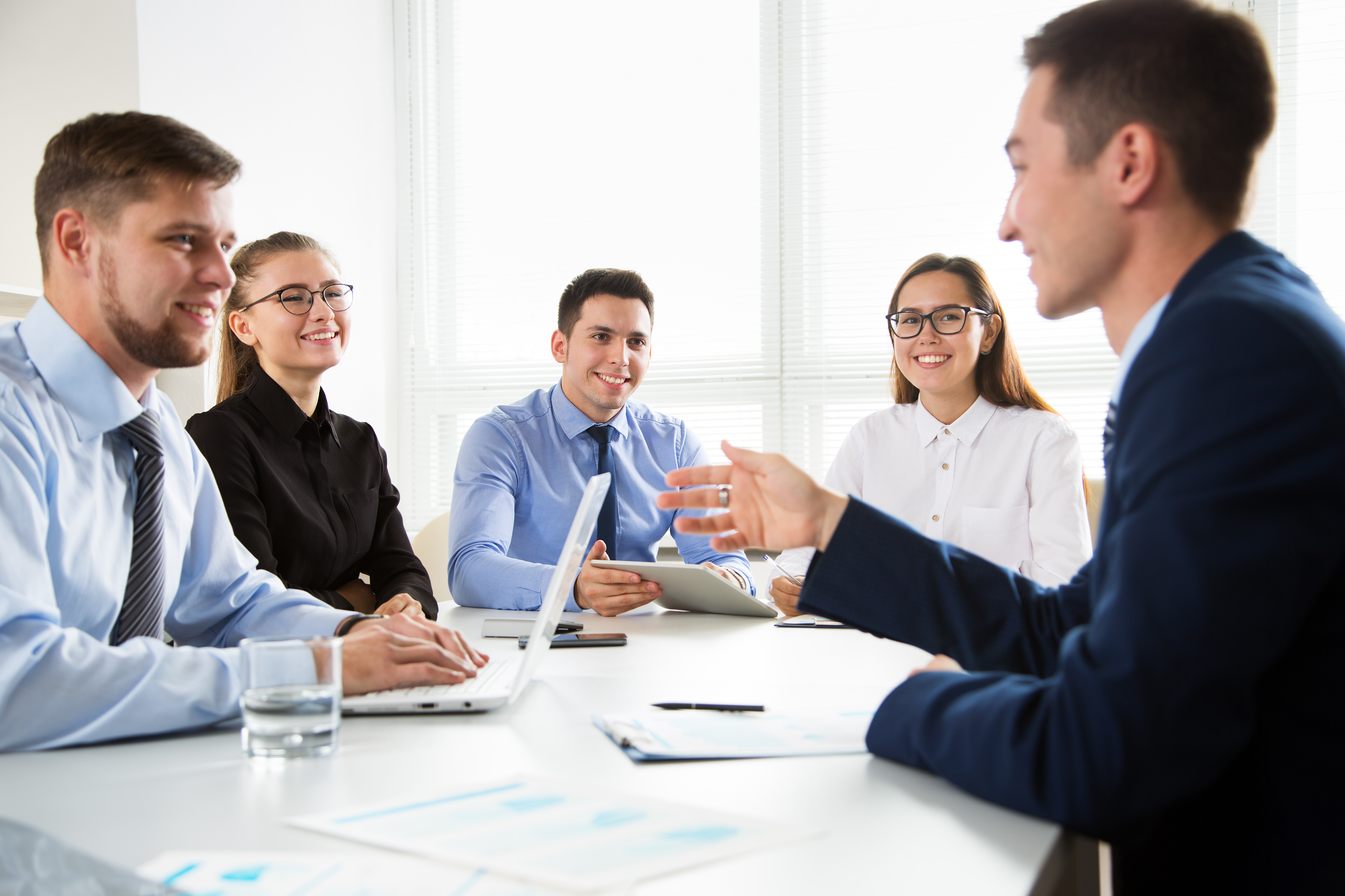 A diverse group of five professionals engaged in a business meeting, sitting around a conference table with laptops and documents in a well-lit office